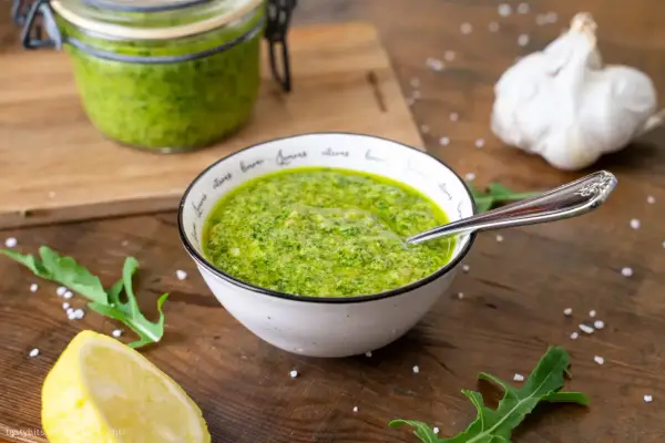 Frisches Rucola Pesto in einer Schüssel, daneben Zitrone, Knoblauch und Rucolablätter.